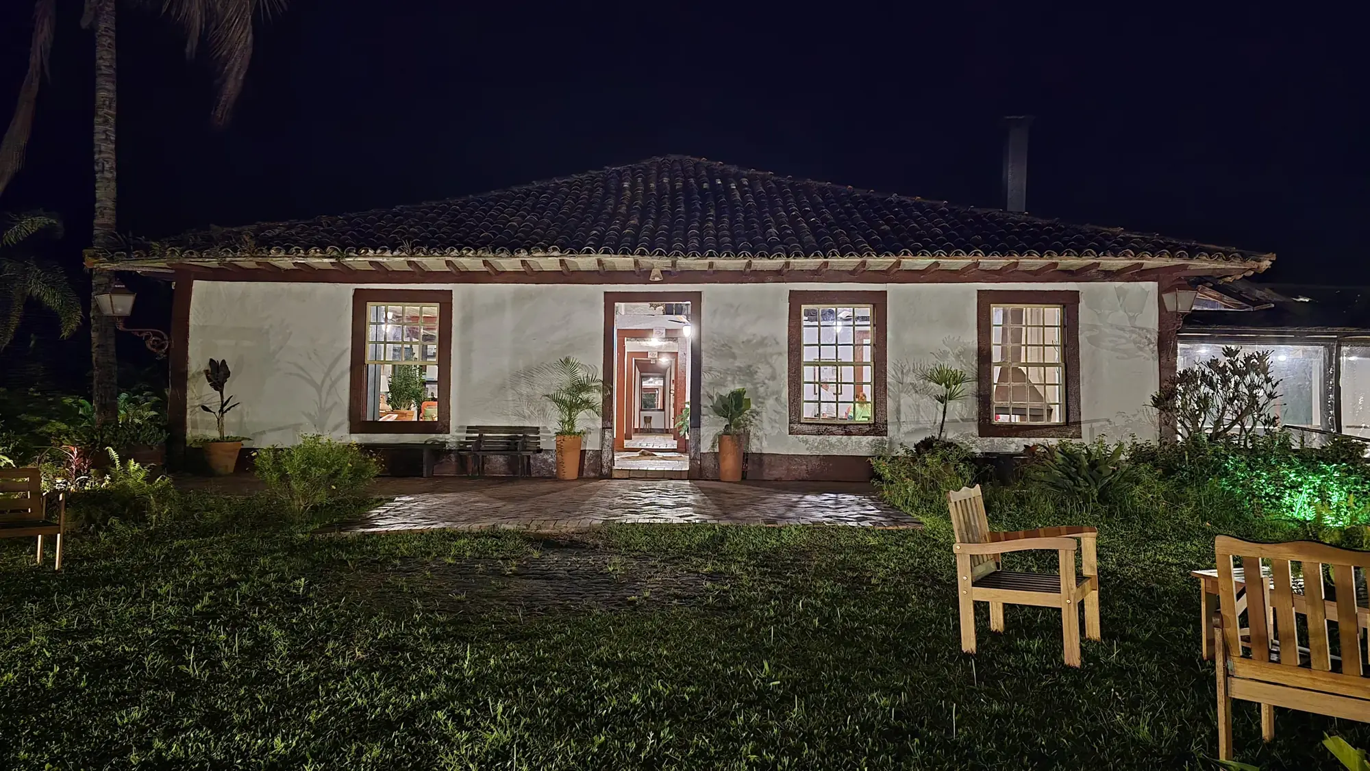 Casa colonial da fazenda iluminada à noite, com o gramado e cadeiras de madeira