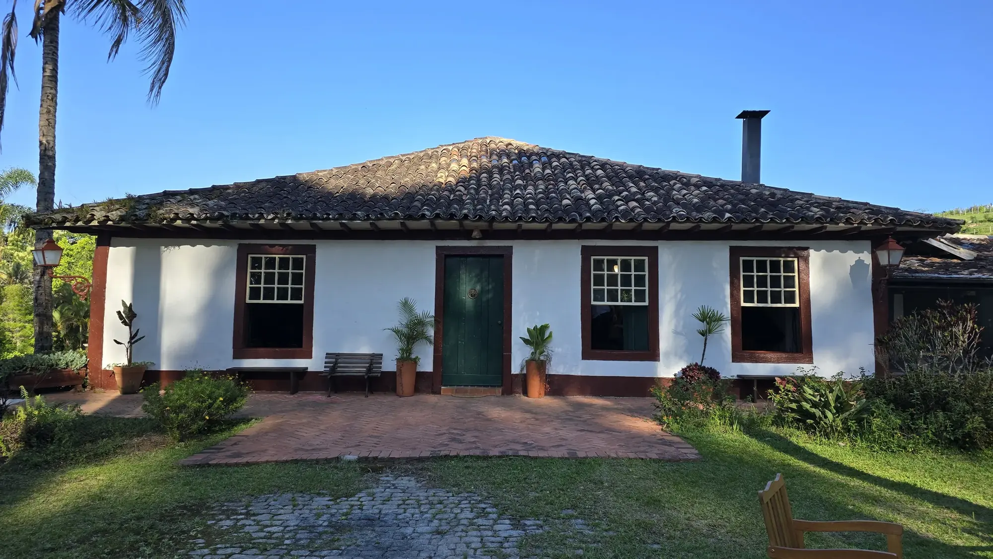 Casa colonial branca com janelas e porta verde, telhado de barro e jardim da fazenda