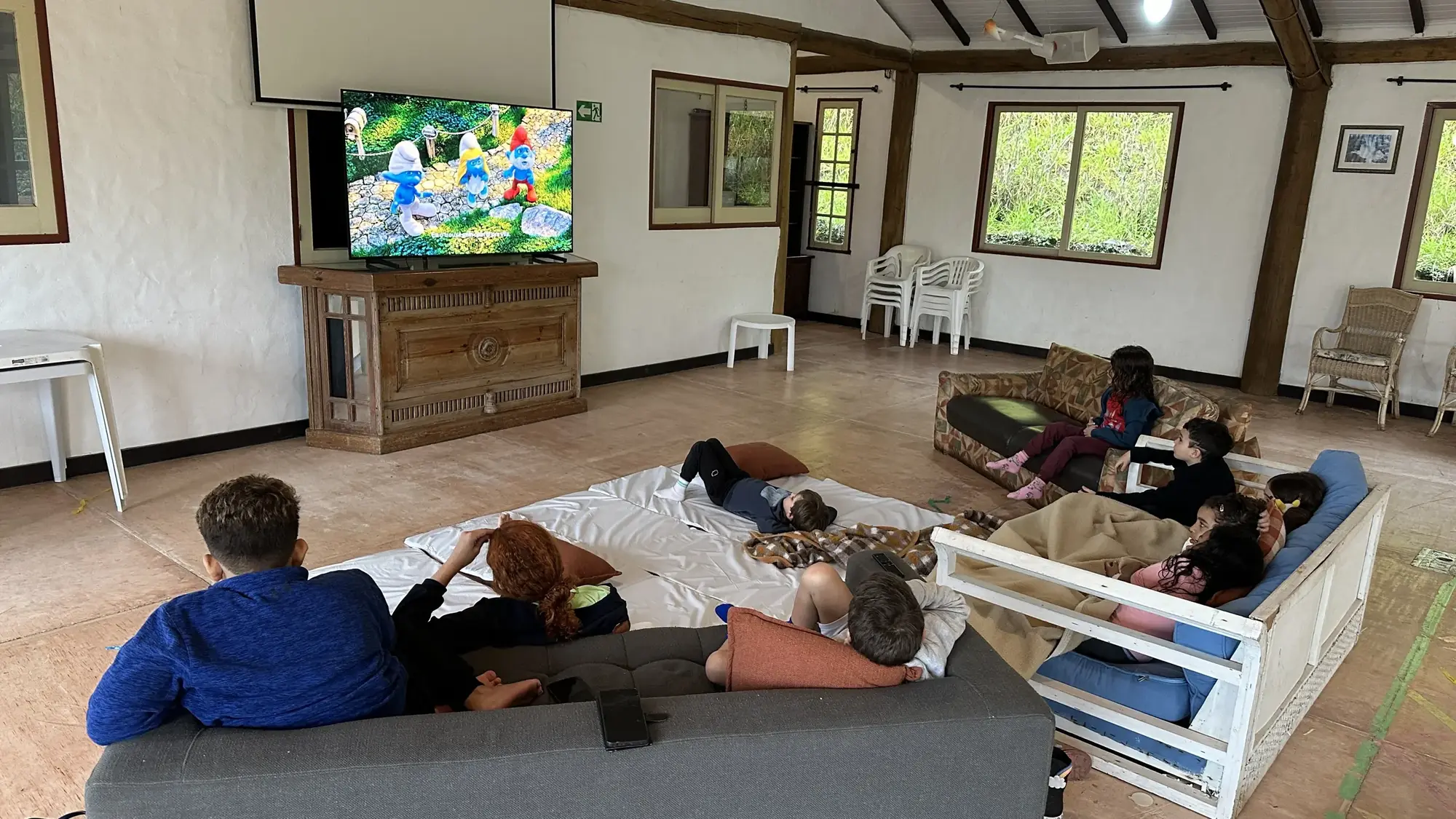 Crianças deitadas nos sofás e colchões assistindo desenho no salão da fazenda