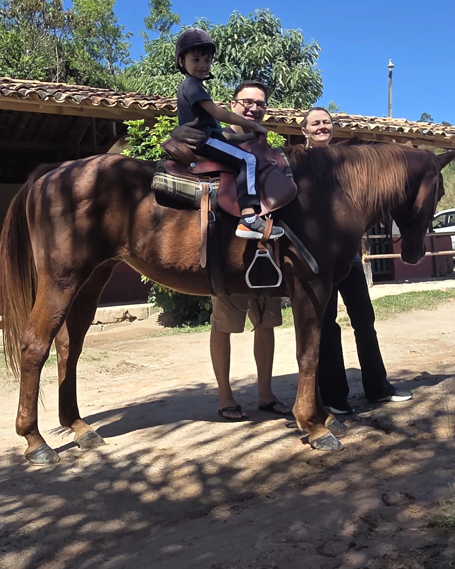 Menino de capacete montado em um cavalo marrom em frente às baias da fazenda
